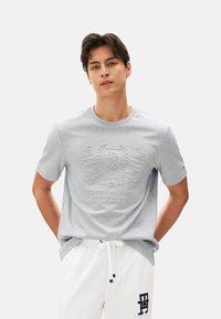 Camiseta corta de manga con logo embossado en el pecho; de color gris claro; hecha de suave tejido de algodón; diseño de cuello redondo.