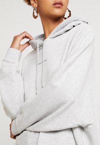Jasnoszara bluza z kapturem, wykonana z miękkiego materiału, z krojem zakładanym przez głowę, sznurkiem ściągającym i ściągaczami na rękawach. Posiada małe logo na klatce piersiowej.