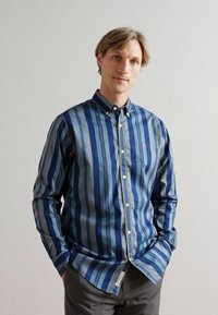 Srajca na gumb z navpičnimi modrimi in sivimi črtami. Narejena iz bombaža, s ovratnikom in gumbastim zapiranjem, teksturirana tkanina in detajl logotipa.