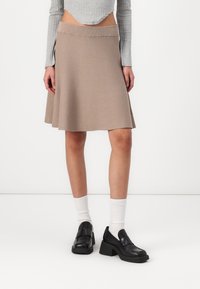 Jupe évasée beige avec une texture tricotée lisse, comportant une large ceinture et un ourlet qui atteint le milieu de la cuisse. Portée avec des chaussures noires et des chaussettes blanches.
