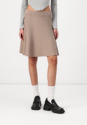 Jupe évasée beige avec une texture tricotée lisse, comportant une large ceinture et un ourlet qui atteint le milieu de la cuisse. Portée avec des chaussures noires et des chaussettes blanches.