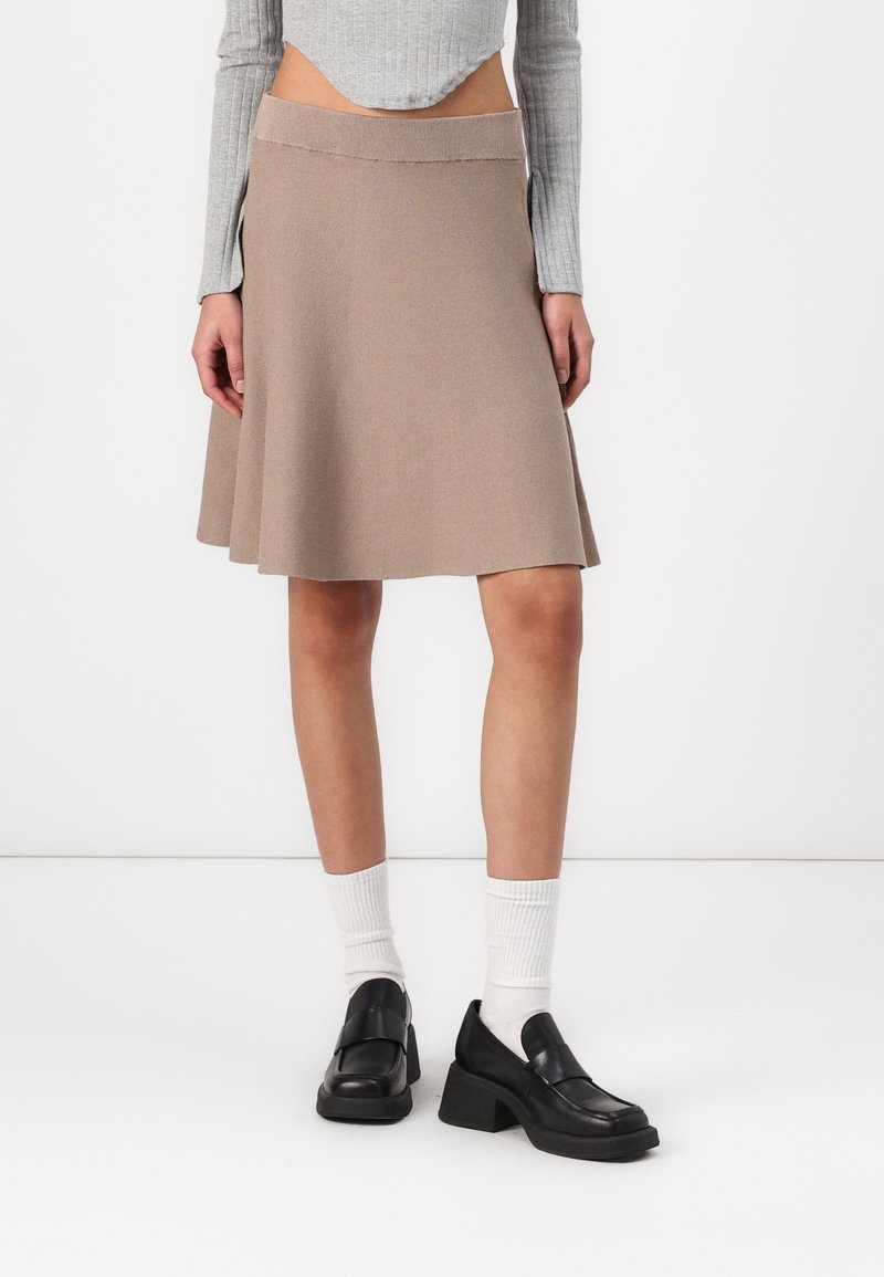 Jupe évasée beige avec une texture tricotée lisse, comportant une large ceinture et un ourlet qui atteint le milieu de la cuisse. Portée avec des chaussures noires et des chaussettes blanches.