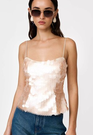 Žena v veľkých slnečných okuliaroch, béžovom topu s flitrami s tenkými ramienkami a modrých džínsach, stojaca na svetlom pozadí.