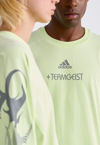 Svijetlozeleni pamučni majici s okruglim izrezom. Jedna ima grafički dizajn, dok druga prikazuje "adidas +TEAMGEIST" u sivom tisku.