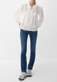 Persona che indossa una blusa bianca a maniche lunghe con pannelli in pizzo, jeans slim-fit blu scuro e sneakers chunky bianche, in piedi con le mani sui fianchi.
