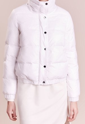 Vrouw draagt een witte donzen jas met zwarte drukknopen over een witte rok, staand tegen een beige achtergrond.