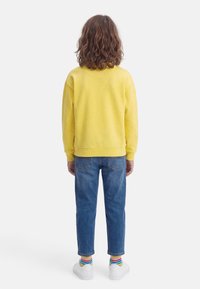 Παιδί με καστανά μαλλιά μέχρι τους ώμους, που φοράει κίτρινο φούτερ, μπλε τζιν, ριγέ κάλτσες και λευκά αθλητικά παπούτσια, στεκόμενο με την πλάτη προς τα έξω.