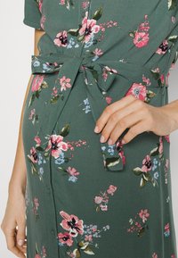 Abito floreale in tessuto verde scuro, con fiori vivaci nei toni del rosa, blu e bianco. Presenta una vita con nastrino e chiusura a bottoni. Motivo testurizzato.