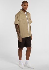 Beige short-sleeve shirt with two chest pockets, worn over a white t-shirt, paired with black shorts and white sneakers.