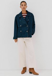 Dżinsowa kurtka zapinana na podwójne guziki w ciemnoniebieskim kolorze z wyraźnymi klapami, srebrnymi guzikami i luźnym krojem, zestawiona z kremowymi spodniami typu wide-leg i brązowymi botkami za kostkę.