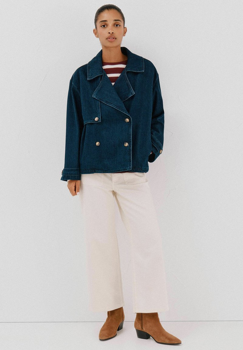 Dżinsowa kurtka zapinana na podwójne guziki w ciemnoniebieskim kolorze z wyraźnymi klapami, srebrnymi guzikami i luźnym krojem, zestawiona z kremowymi spodniami typu wide-leg i brązowymi botkami za kostkę.