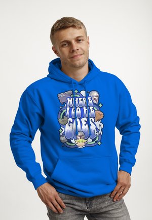 Ung mand iført en lys blå hættetrøje med teksten "Where I Go He Goes" og Star Wars Mandalorian-tema illustrationer på fronten, smilende let.