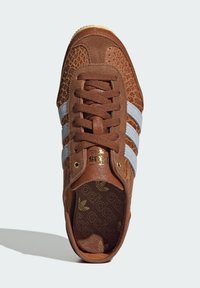 Brązowy zamszowy sneaker z teksturowaną cholewką, szarymi skórzanymi paskami i sznurowanym zapięciem. Złote logo widoczne na języku i wkładce.