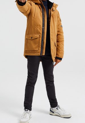 Personne portant une doudoune beige, un pantalon noir et des baskets blanches, debout avec un bras levé, visage non visible.