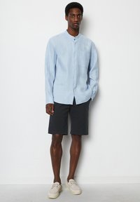 Lichtblauw linnen shirt met een bandkraag en borstzak, gecombineerd met donkere shorts en lichtgekleurde schoenen, staand tegen een witte achtergrond.