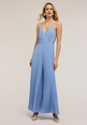 Kvinde iført en lyseblå plisseret buksedragt med tynde stropper, hvide spidse sko, guldringøreringe og lag-på-lag halskæder mod en ensfarvet baggrund.