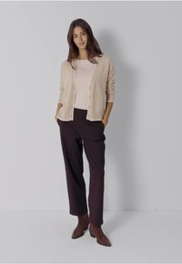 Beżowy, dziany kardigan z guzikami, noszony na jasnej bluzce, zestawiony z ciemnobrązowymi spodniami i brązowymi botkami za kostkę. Gładka faktura w całym ubraniu.