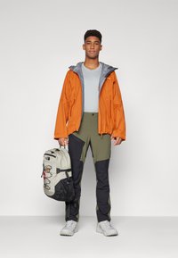 Veste imperméable orange avec des accents gris, t-shirt gris, pantalon olive et noir, tenant un sac à dos beige avec des accents foncés.