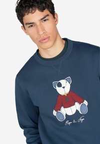 Sudadera azul marino con un gráfico de un oso con un suéter rojo, costuras detalladas y acentos azules. Cuello redondo con tejido texturizado.