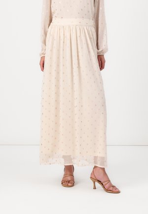 Kvinde iført en lang, lys beige nederdel med små metaliske prikker og beige sandaletter med tynde stropper, stående på et hvidt gulv.