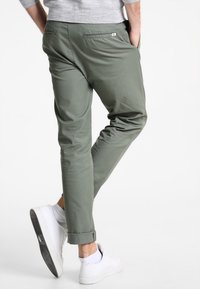 Muž v olivově zelených slim-fit kalhotách s ohrnutými nohavicemi, šedém svetru a bílých teniskách, stojící s rukama v kapsách.
