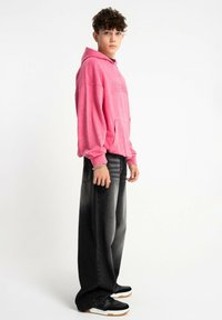 Różowa bluza z kapturem z kieszenią z przodu, zestawiona z czarnymi szerokimi spodniami dżinsowymi z gradientowym efektem. Uzupełniona czarnymi trampkami.