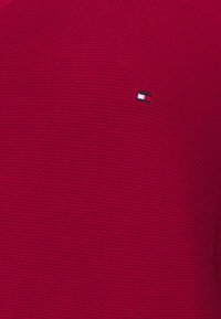 Pull en tricot rouge avec une texture côtelée ; présente un petit logo en bleu, blanc et noir sur la partie gauche de la poitrine.