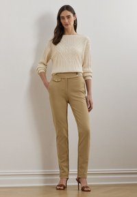 Γυναίκα που φοράει κρέμ του πουλόβερ με πλεξούδα, καφέ παντελόνι ψηλής μέσης με λεπτομέρειες από κουμπιά και καφέ σανδάλια με τακούνι, που στέκεται μπροστά σε έναν απλό τοίχο.