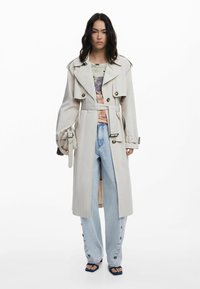 Light beige trench coat with wide lapels, double-breasted buttons, and a belted waist. Worn over a patterned top and light blue wide-leg jeans.
