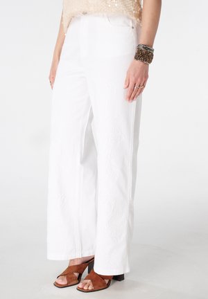 Femme portant un pantalon blanc à jambes larges avec une broderie florale subtile, des sandales à talons couleur cuir clair, un top beige à sequins et des bracelets empilés au poignet droit.