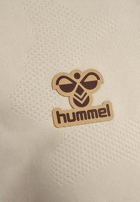 Cremfarbener Stoff mit einem strukturierten Muster, versehen mit einem braunen, geprägten Bienenlogo und dem Wort "hummel" in fetter Schrift.
