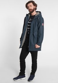 Námořnická voděodolná bunda se šedým fleecovým podšívkou, nastavitelnou kapucí, předním zipem a postranními kapsami, nošená přes černé tričko s potiskem a černé kalhoty.