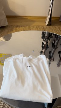 Λευκό βαμβακερό T-shirt με στρογγυλή λαιμόκοψη, με μικρό μαύρο λογότυπο Nike στο εμπρόσθιο μέρος, τακτοποιημένο διπλωμένο πάνω σε μια ανακλαστική επιφάνεια.