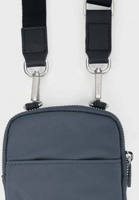 Sac bandoulière en nylon gris avec une texture lisse, fermeture zippée et deux bretelles réglables. Accents en métal argenté et détails cousus visibles.