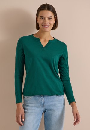 Femme aux cheveux courts bruns souriante, portant un haut vert à manches longues avec un encolure en V et un jean bleu clair, debout contre un mur beige.