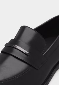 Mocassin en cuir noir à fini lisse, avec une lanière décorative portant une petite plaque métallique avec logo, et un design à bout arrondi.