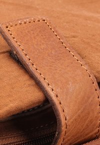 Brun læderrem med synlig syning, fastgjort til en struktureret stofbaggrund. Nærbillede fremhæver materialer og håndværksdetaljer.