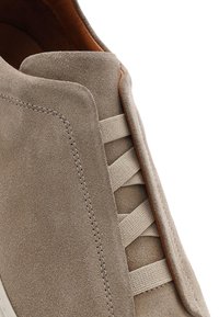 Beżowy zamszowy but wsuwany z elastycznym sznurowaniem i brązową skórzaną wyściółką wewnętrzną, zbliżenie na górną część i detale szwów.