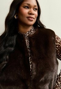 Brązowa kamizelka z futra syntetycznego o puszystej fakturze, noszona na białej i brązowej bluzce w panterkę. Lekko zaokrąglona linia dekoltu, brak widocznych zapięć.