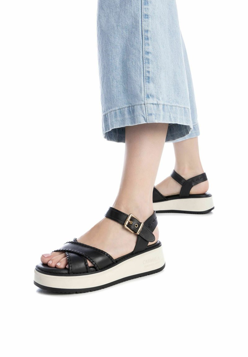 Sandalias de plataforma de cuero negro con un diseño trenzado, correa de tobillo ajustable y suela blanca en contraste. Acabado texturizado y detalles en dorado.