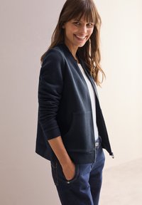 Giacca bomber trapuntata navy con un motivo texturizzato a spina di pesce, polsini a coste e tasche frontali, abbinata a una maglietta bianca e jeans scuri.