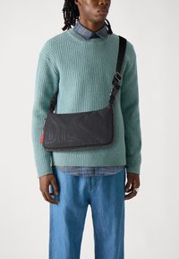 Black fabric shoulder bag with embossed "DIESEL" logo, silver zipper, red tag detail. Worn over a light blue ribbed sweater.
