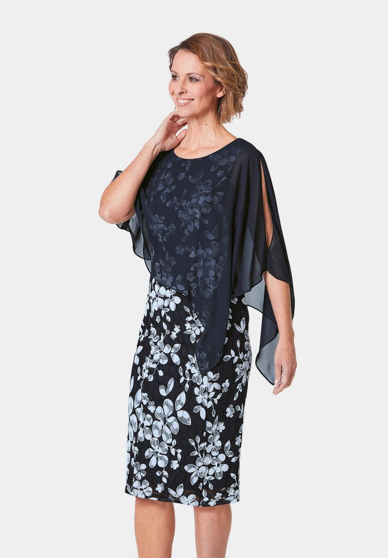 Blumenkleid mit marineblauem Grund, weißem Blumenmuster und einem fließenden Chiffon-Überzug mit offenen Ärmeln, der einen geschichteten Effekt erzeugt.