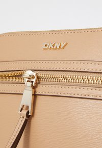 Primo piano di una borsa beige DKNY con hardware dorato, cerniera visibile e materiale in pelle testurizzata. Presenta un dettaglio con logo impresso.