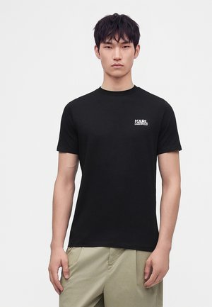 Ung mand med kort sort hår iført en sort Karl Lagerfeld t-shirt og beige bukser, står mod en ensfarvet lys baggrund.