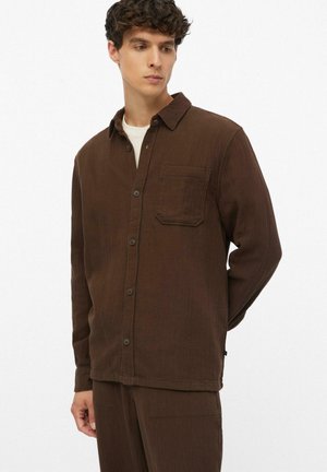Junger Mann trägt ein braunes Hemd mit Brusttasche und passende braune Hose, steht mit den Händen hinter dem Rücken.