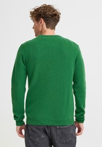 Zielony sweter dziany, z okrągłym dekoltem, długimi rękawami, prążkowanymi mankietami i pasem, miękka faktura, noszony z szarymi dżinsami.