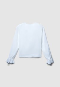Blusa bianca a maniche lunghe con polsini arricciati e scollatura rotonda, esposta in piano su uno sfondo grigio chiaro.