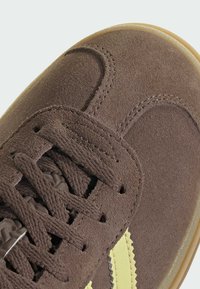 Brune semskede sneakers med en teksturert finish, med gule striper, runde snørelisser og en gummisåle. Detaljene inkluderer sømdekorer.