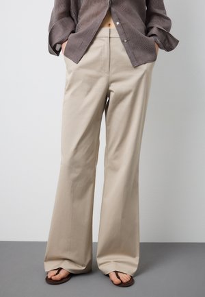 Persona con pantalones anchos beige, camisa marrón a rayas parcialmente metida, con las manos en los bolsillos y sandalias marrones sobre suelo gris.